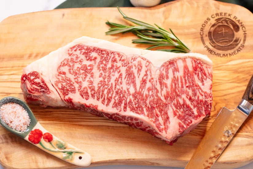 A American Wagyu New York Strip Steak on a wooden cutting board, accompanied by a steak knife, salt, pepper, and garlic, with a logo stamped on the lower right corner of the image.